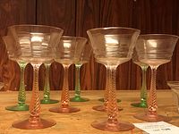 Ten vintage stemmed glasses with twisted green and pink stems, arranged with a wood panel background.