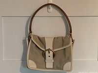 Front view of beige canvas shoulder bag with white leather trim, gold-tone buckle closure, and brown leather handle.