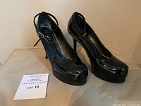 Front-side angled view of the black patent leather platform pumps with ankle strap, showing rounded toe and glossy appearance.