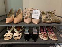 Six pairs of women's shoes on metal shelving: nude pointed toe pumps, nude strappy pumps, gold open toe sandals, black kitten heel mules, cream flower motif pumps, and white sandals with red stitching.