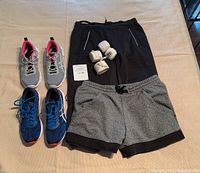 Photo showing blue Asics Gel DS Trainer shoes, grey and pink Asics road running shoes, grey shorts, black pants, and white boxing wraps laid out on beige surface.