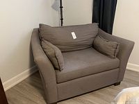 Gray fabric chair and a half with two matching rectangular pillows, positioned in corner of room with wooden floor and dark curtain.