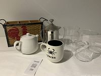 Photo showing woven cutlery holder, white ceramic teapot, ceramic mug with Cancer zodiac sign, stainless steel milk frother, and two clear ribbed glass cups on a table.