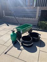 Photo showing a green broadcast spreader, green handheld sprayer, and multiple coils of black plastic lawn edging strips on a paved surface in daylight.