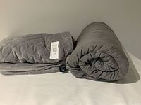 Full view of folded grey plush cover and rolled grey weighted blanket on a white surface.