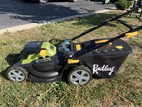 Side view of Radley battery powered lawnmower on grass showing 16 inch cutting width, black and green body with yellow adjustment levers and a black grass catcher bag with Radley logo.