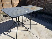 Two metal outdoor bar height tables outdoors on concrete against wooden fence. Tables have square slatted tops with umbrella holes, one table surface shows rust and discoloration.