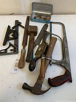 Nine vintage hand tools including hammer, saw, mallet, rasp, brace, plane, corner braces, and a small metal box with hardware on a white surface.