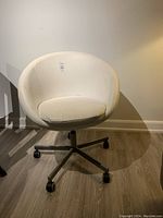 White curved office chair with padded seat cushion, metal base, and rolling casters shown in room.