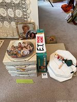 Annie collector plates stacked in boxes with vintage memorabilia including framed photos, E.T. phone home item, and a decorative figurine, on a carpeted floor.