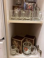 Six clear glass mugs with A&W logos on a shelf with boxed mugs below.