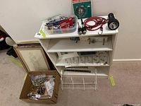 Photo showing white bookshelf with framed art leaning against it and box of miscellaneous hardware including metal hinges and fittings.