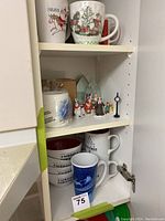 Photo of white ceramic mugs and bowls with Christmas text and graphics along with small holiday figurines inside a cupboard.