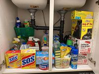 Full view inside under-sink cabinet showing assorted cleaning supplies, sponges, garbage bags, and spray bottles arranged on shelves.