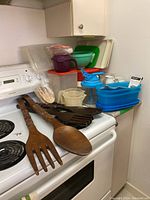Photo showing large wooden spoon and fork set laid on stove with plastic storage containers and measuring cups stacked behind.