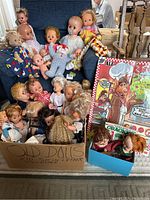 Box and chair containing multiple assorted vintage dolls of various sizes and materials. Dolls show signs of age and wear.