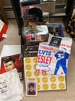 Photo showing Elvis memorabilia items including a boxed collector's record, a white Elvis t-shirt with print, a framed mirror with Elvis silhouette, vintage magazines, and collectible plate.