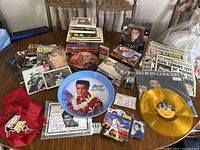 Photo of assorted Elvis Presley books, magazines, CDs, cassette tapes, vinyl records including a yellow translucent record, and commemorative plate with Elvis image on a table.