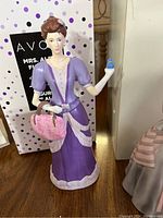 Porcelain figurine of a woman in a purple dress holding a blue bottle and pink handbag with Avon themed box behind.