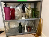 Wide view of metal shelving unit holding a variety of glass vases and containers in different shapes, sizes, and colors including a large clear bowl, green bottles, yellow tall vase, and others