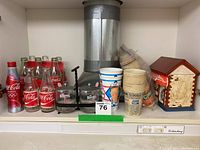 Image showing assortment of vintage Coca Cola bottles, aluminum Coca Cola Olympic bottle, Fields cookie jar shaped like a house, assorted bar items, plastic coasters, and a metal beverage dispenser with a pump.