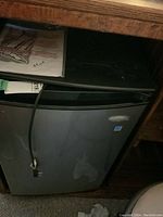 Front view of the Whirlpool black bar fridge with visible plug cord and manual on top.