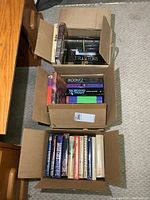View of three open boxes filled with assorted books including Harry Potter and multiple novels.