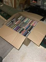 Top view of large cardboard box filled with stacked music CDs, showing the large quantity of CDs packed inside.