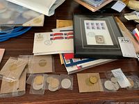 Overview of various collectibles including coins in cardboard sleeves and plastic holders, framed stamp sheet, and loose stamp pages on table.