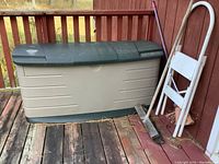 Rubbermaid plastic deck box with dark green lid, beige body, placed on wooden deck next to red wall, with broom and ladder nearby.