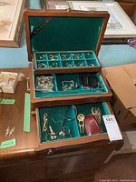 Open wooden jewelry box with three tiers filled with assorted jewelry including rings, earrings, necklaces, and small accessories on teal velvet lining.