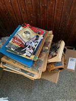Photo showing stacked sports magazines, loose papers, and boxed ephemera including a baseball in a display case.