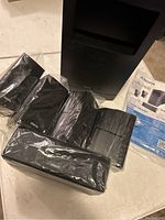 Photo showing five smaller Alexa Acoustic speakers wrapped in plastic, a large subwoofer, and instruction manual on a white surface.