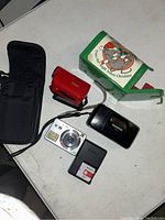 Photo showing three cameras and accessories on a white surface including a silver Sony Cyber-shot camera, black Olympus camera, red Vivitar camera, black battery charger, and a soft black carrying case.
