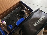 Top view of box showing large black brush attachment, blue and black parts, and small white spray bottle.