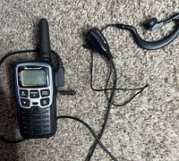 Photo of Midland walkie talkie and wired earpiece on carpeted surface.