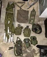 Military bed frame and assorted army accessories arranged on floor, including bed frame, medical bag, canteens, pouches, belt, and helmet cover.