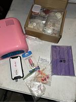 Photo shows pink nail UV lamp, boxed nail tips, pedicure set, cuticle oil, and nail decorations on a surface.