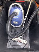 Photo showing blue Oreck vacuum canister inside a black plastic bin with hose attached, metal wand, and power nozzle placed coiled in the bin.