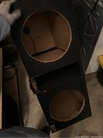 Photo showing angled view of the black carpet-covered speaker box with two circular subwoofer cutouts and rectangular bass port in between.