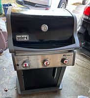 Front view of the Weber Spirit gas grill showing black lid with thermometer, stainless control panel with three control knobs, and lower cabinet.