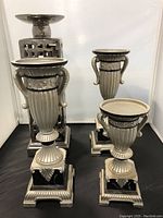 Photo showing four silver-colored decorative candleholders and one fruit bowl with detailed ornate designs and black bases.