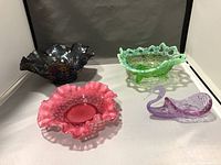Four carnival glass pieces displayed: three ruffled edge bowls in dark iridescent, green, and pink colors, plus one purple glass swan figurine.