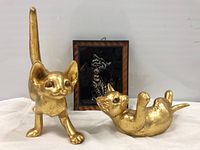 Two gold leaf pottery cat sculptures and one framed cat etching on display against a white backdrop.