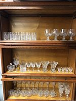 Full view of three shelves with assorted types of crystal and cut glass drinkware arranged on wooden shelves.