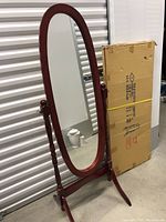 Assembled oval floor mirror standing beside unopened boxed mirror in storage setting