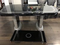 Front angle view showing table top with black glass surface and silver Greek-key decorative border, dual rounded pedestal bases with silver vertical stripes