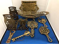 Overview photo of 10 brass Judaica items laid on blue fabric including tray, basket, trivets, napkin holders, bottle opener, and key