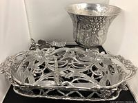 Multiple pewter and silverplate serving trays with floral and lattice designs, champagne bucket with grapevine embossing, and other serving accessories in a black background white-walled setup.