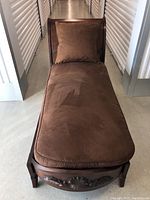 Full view of chaise longue showing brown upholstered cushion, wood frame, wicker back, and carved foot panel